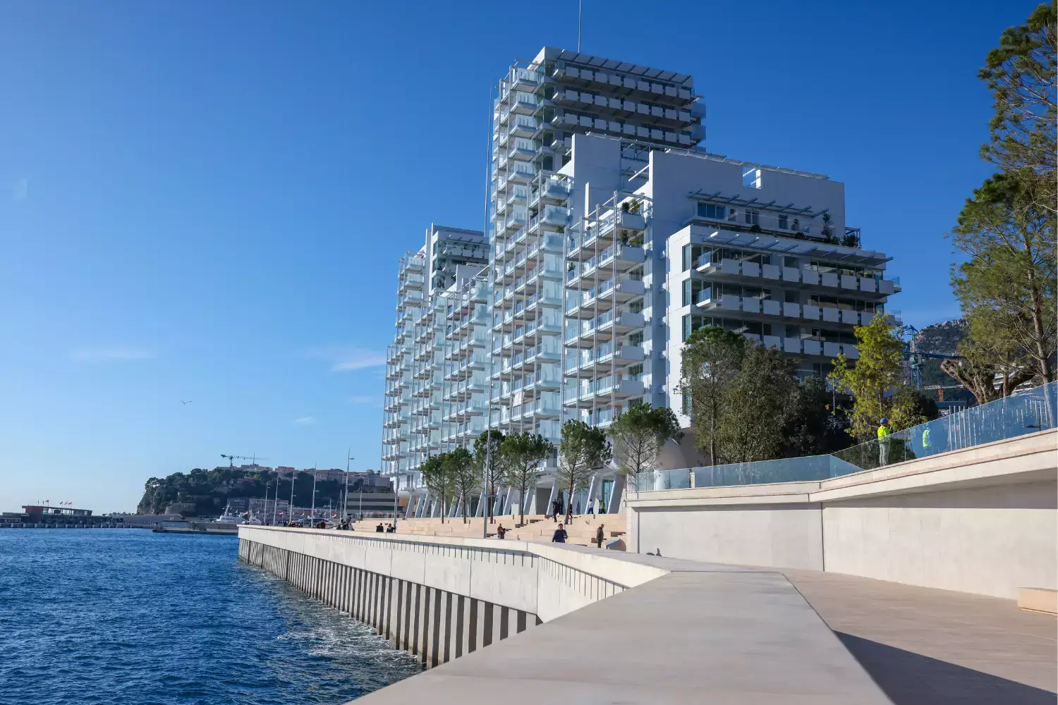 Le Renzo, a multi-tiered white residential building with glass balconies rising above the Mareterra waterfront promenade under a clear blue sky